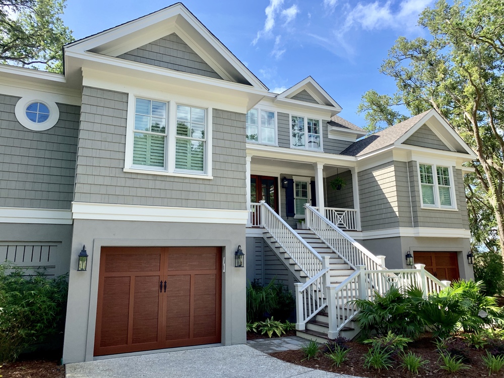 A three-story home with light gray fiber cement cedar shake and two wooden garage doors. There is a large stairway leading to the front door.