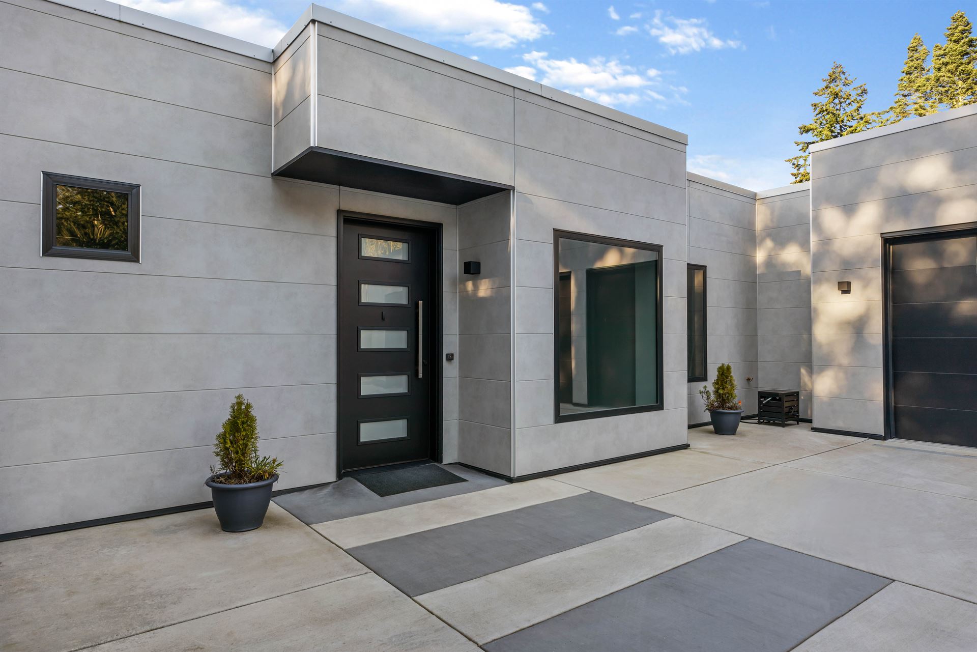 The front entrance of a very modern home, featuring a black front door, black garage door and black framed windows surrounded by light gray wide panel fiber cement siding.