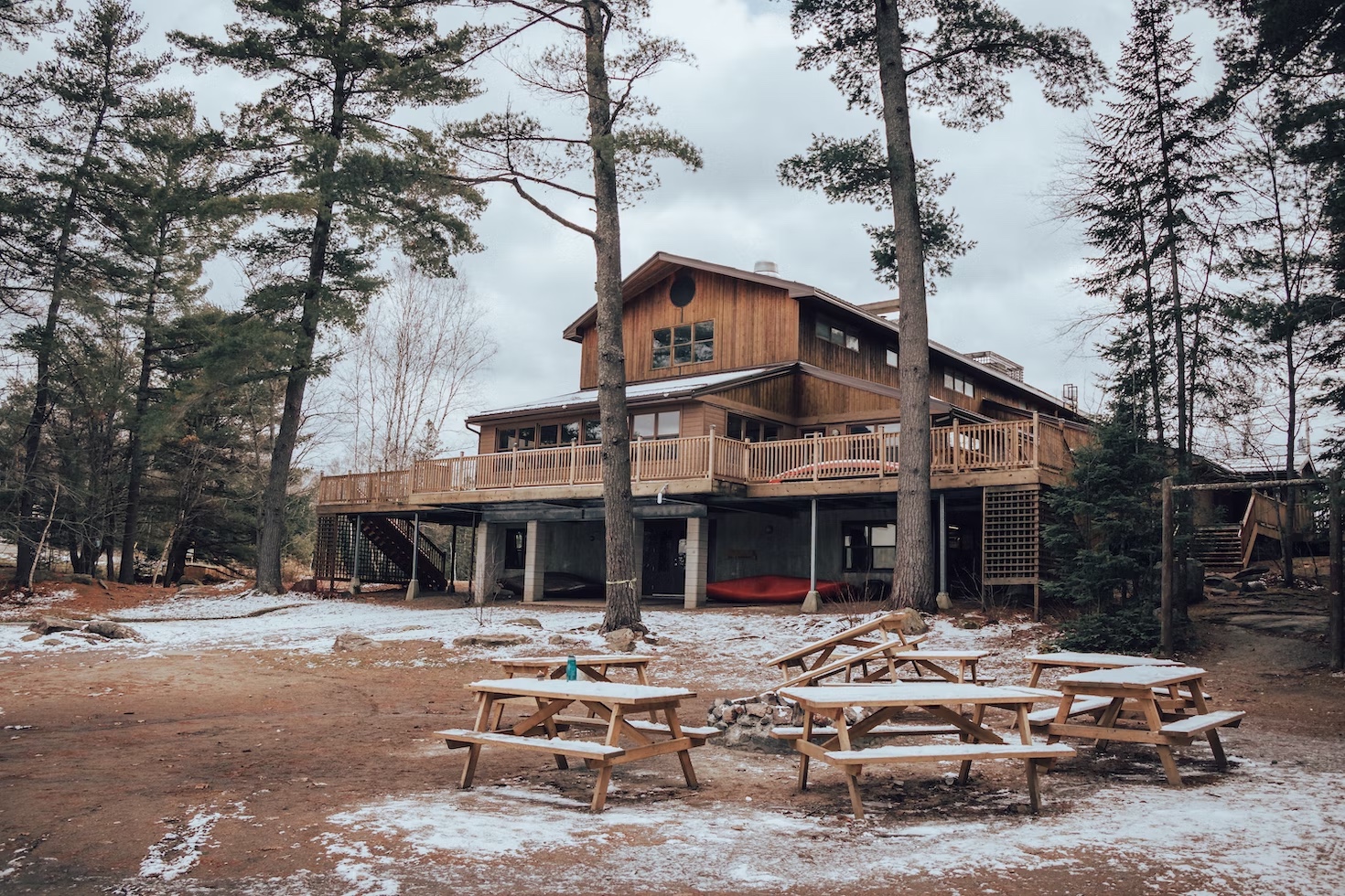 The back deck of a three-story home with wood siding surrounded by trees. Several picnic tables sit in the backyard lightly covered in snow.