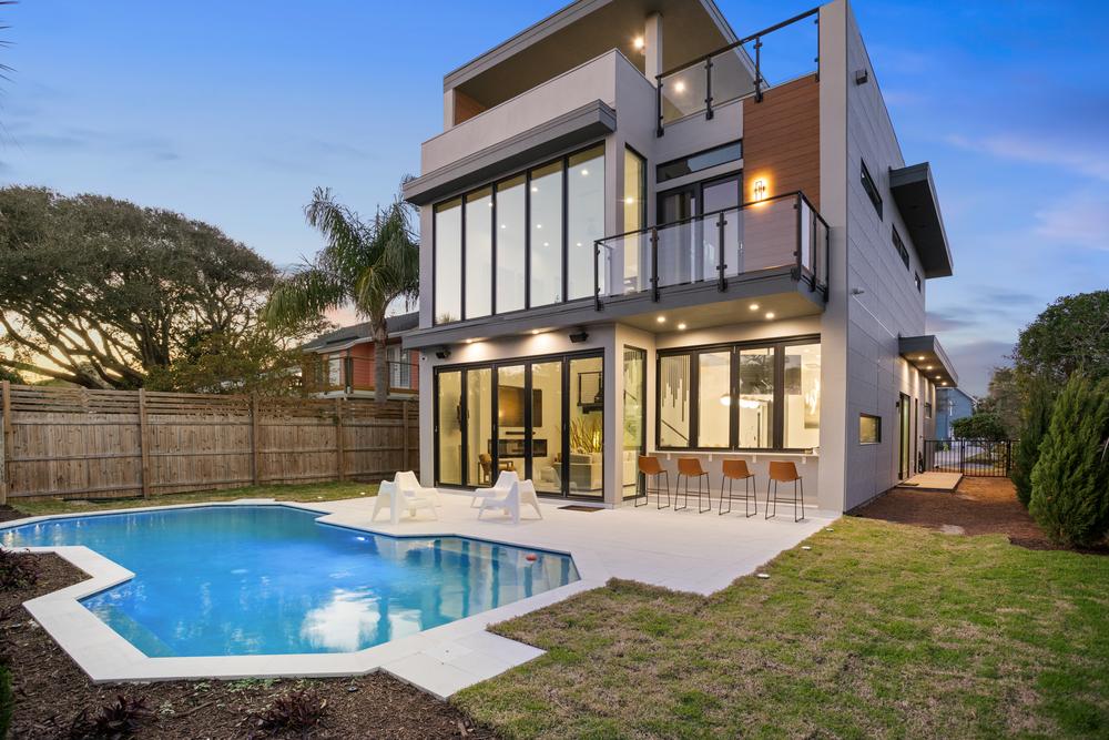 A three-story modern home has a pool in the backyard. There is an outdoor bar area, and the home has large glass windows that overlook the pool area.