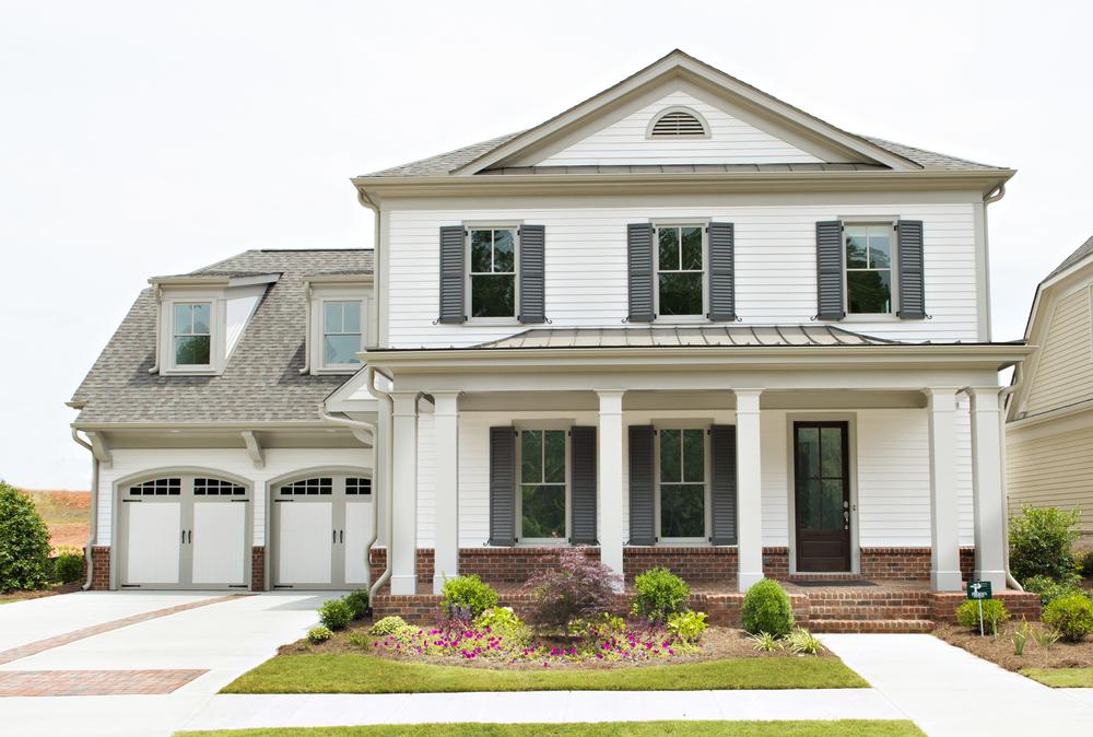 This traditional-style home has white siding, black shutters, a two-car attached garage and a concrete driveway. There is a brick front porch with six pillars. 