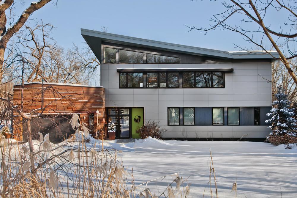 A gray and wooden home with a 45-degree shaped, a flat roof is focused in a snowy landscape.